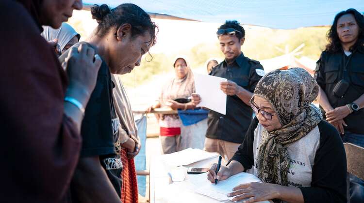 Zwei Frauen stehen vor einem Tisch, dahinter sitzt eine weitere Frau mit einer Liste.
