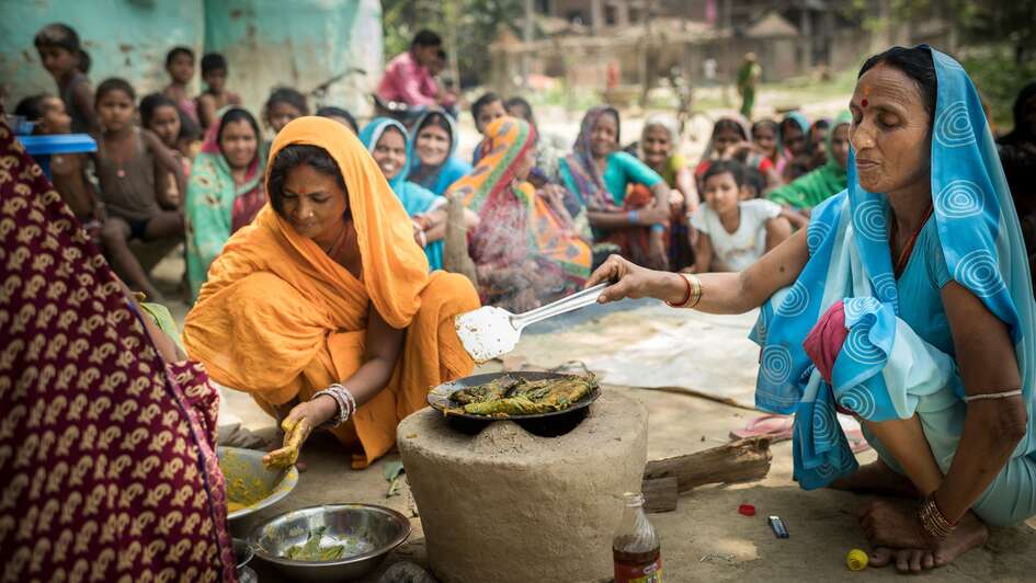 Zwei Frauen kochen, im Hintergrund schauen mehrere Frauen zu.