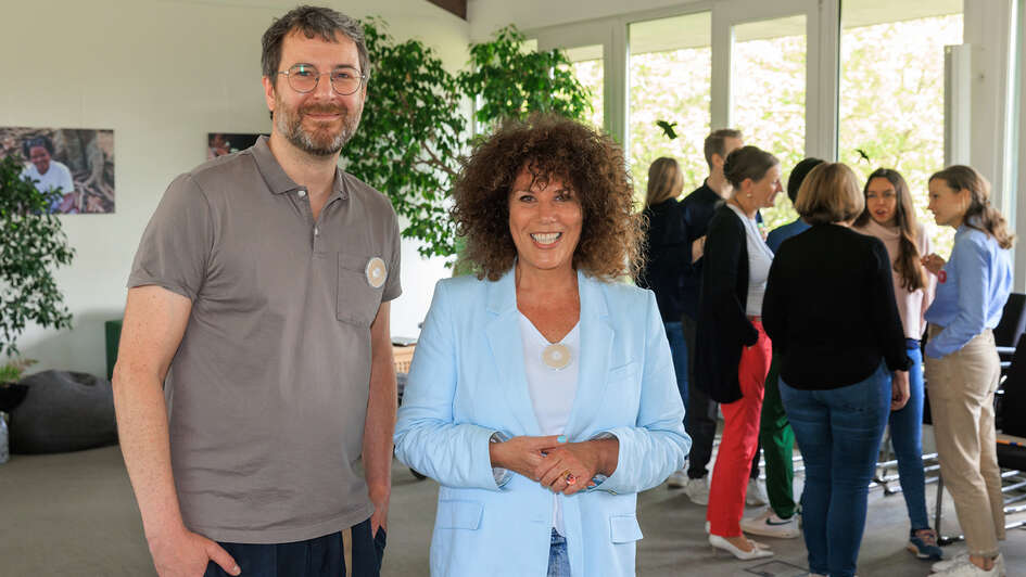 Foto aus einem Konferenzraum der Welthungerhilfe. Ruth Wennemar und Ulrich Schlenker schauen im Vordergrund in die Kamera, im Hintergrund unterhält sich eine Personengruppe.