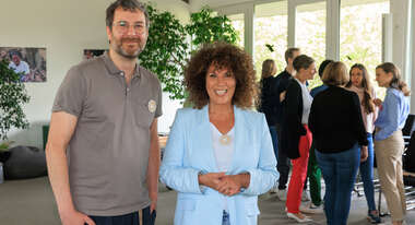 Foto aus einem Konferenzraum der Welthungerhilfe. Ruth Wennemar und Ulrich Schlenker schauen im Vordergrund in die Kamera, im Hintergrund unterhält sich eine Personengruppe.