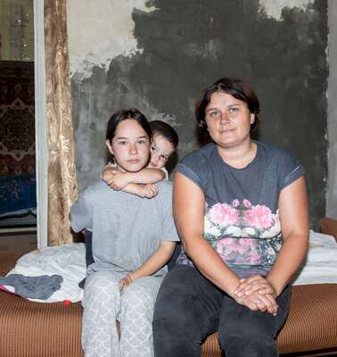 In einer kargen Wohnung sitzen eine Frau und zwei Kinder auf der Kante des Bettes.