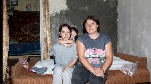 In einer kargen Wohnung sitzen eine Frau und zwei Kinder auf der Kante des Bettes.