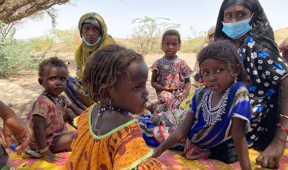 Eine Frau mit Mundschutz und mehrere Kinder sitzen auf einer Decke auf dem Boden.