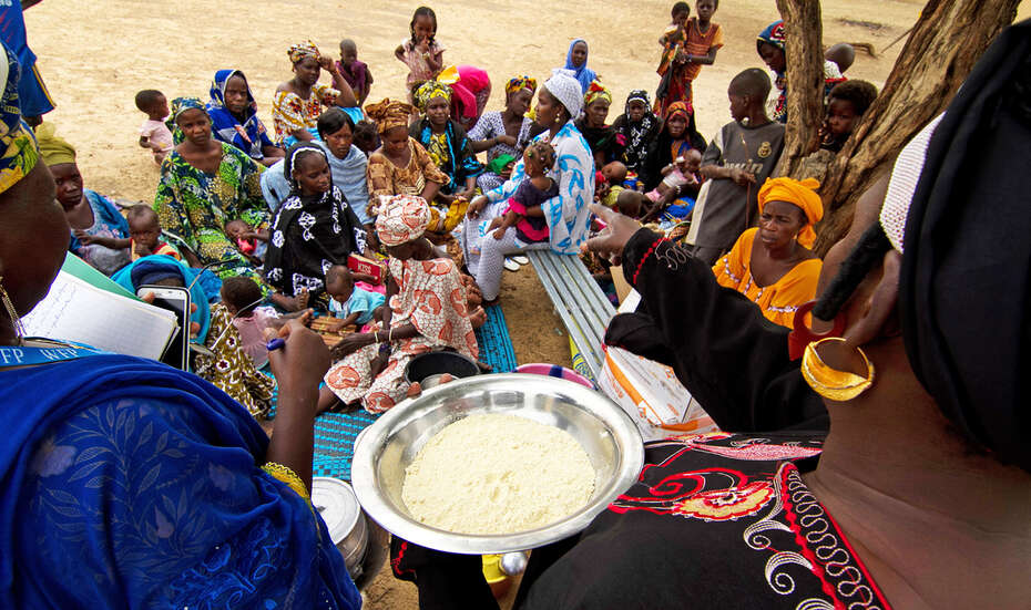 Zwei Trainnerinnen stehen mit einem Teller Brei vor einer sitzenden Gruppe Frauen und Kindern.