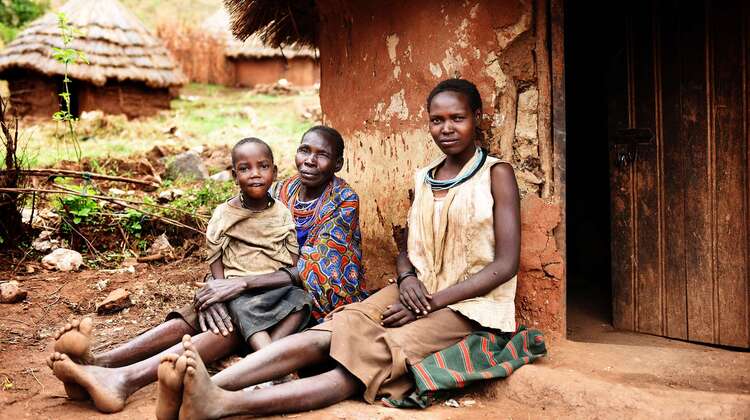 Zwei Frauen und ein Kind sitzen vor einer Hütte und schauen in die Kamera.