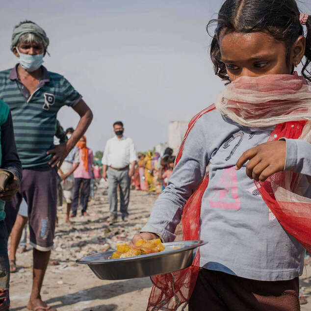Ein Mädchen bedeckt ihren Mund und Nase mit einem Tuch. Sie hält einen Teller mit Essen in der Hand.