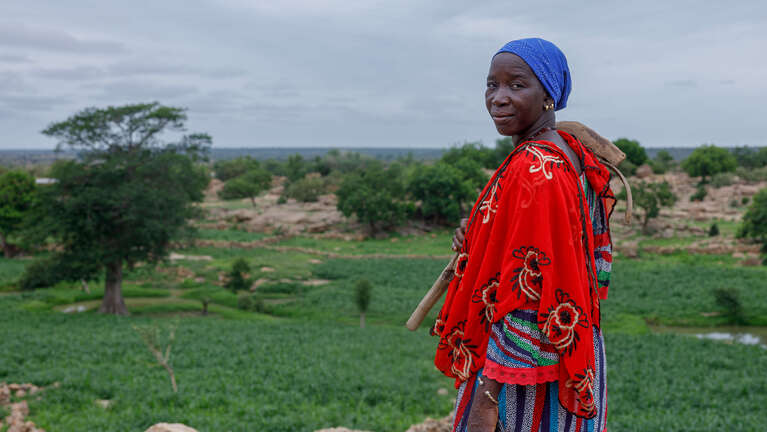 Eine Frau in farbenfroher Kleidung steht mit einem landwirtschaftlichen Werkzeug auf einem Feld in Mali.