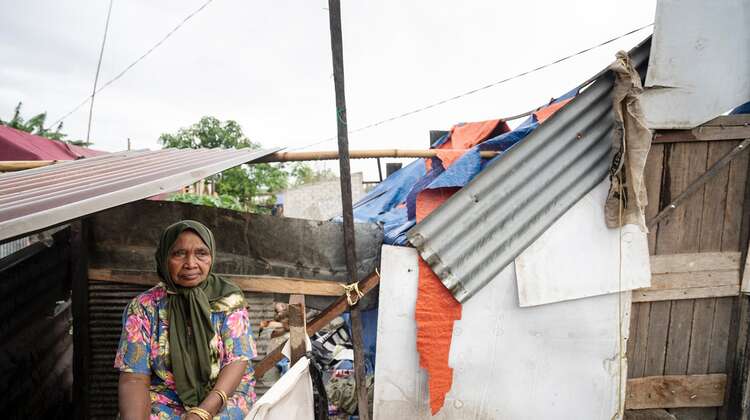 Eine ältere Frau schaut traurig in die Ferne, hinter ihr sieht man ein provisorisches "Zelt" aus Planen und Wellblech.