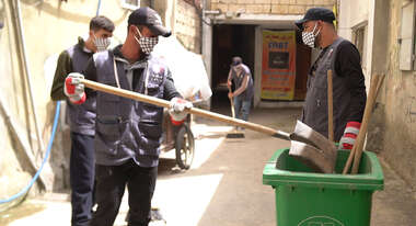 Drei Männer bei der Müllbeseitigung: Ein Mann wirft mit einer großen Schaufel etwas in eine grüne Tonne.