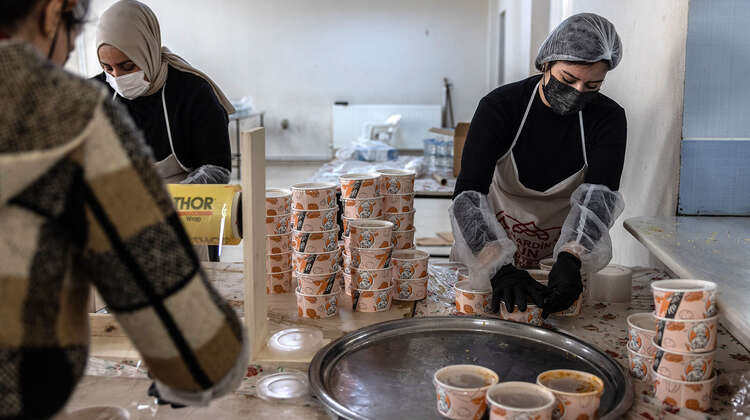 Drei Frauen in Arbeitskleidung verpacken frisch gekochte Mahlzeiten in handliche Transportboxen für die spätere Verteilung.
