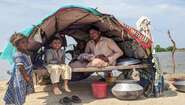 Fluten in Pakistan: Ein Mann, eine Frau, zwei kleine Kinder und ein Baby in ihrer Notunterkunft, die aus Holzstäben, Decken und einer Plastikabdeckung besteht. im Hintergrund ist Wasser zu sehen.