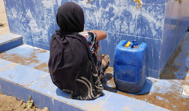 Eine Frau wartet an einer Wasserstelle darauf, dass ihr Kanister mit Trinkwasser voll wird.