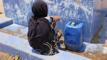 Eine Frau wartet an einer Wasserstelle darauf, dass ihr Kanister mit Trinkwasser voll wird.