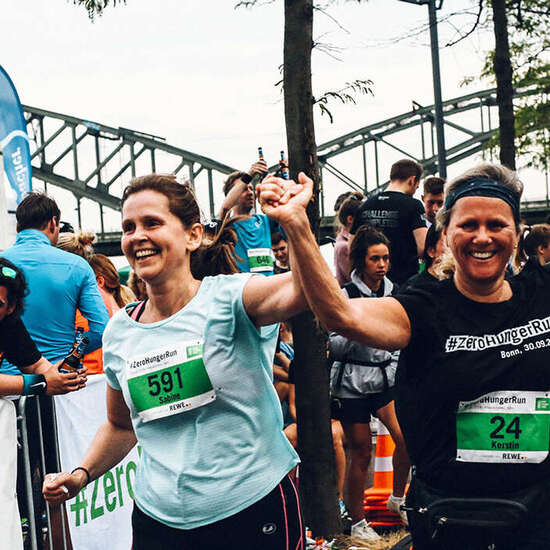Zwei laufende Frauen in Sportkleidung heben jubelnd ihre Arme in die Höhe. Im Hintergrund sind Zuschauer und der Kölner Rheinauhafen zu sehen. 