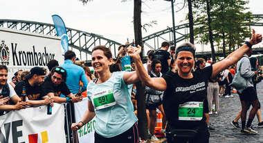 Zwei laufende Frauen in Sportkleidung heben jubelnd ihre Arme in die Höhe. Im Hintergrund sind Zuschauer und der Kölner Rheinauhafen zu sehen. 