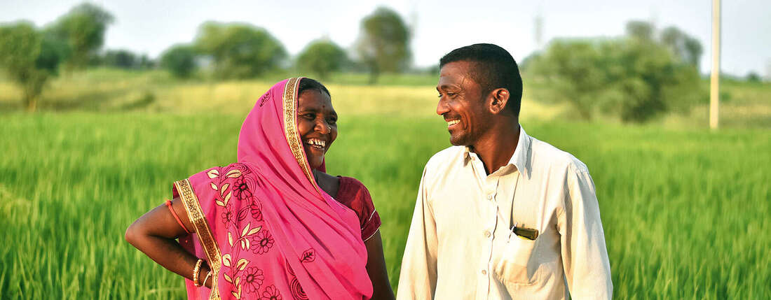 Eine indische Frau und ein indischer Mann stehen in einem grünen Feld und lachen sich an.