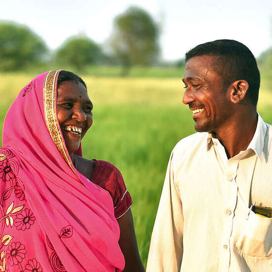 Eine indische Frau und ein indischer Mann stehen in einem grünen Feld und lachen sich an.