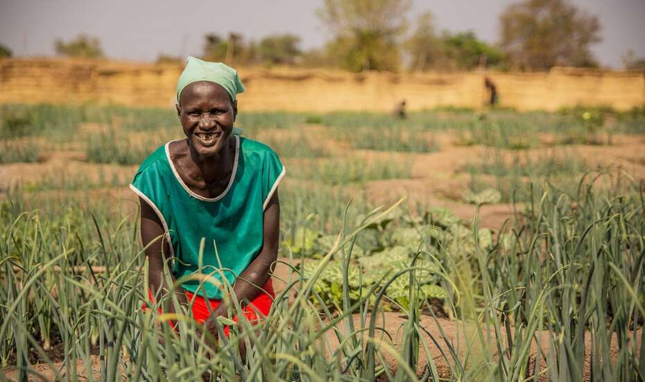 Eine Frau steht in einem Garten und lacht in die Kamera.
