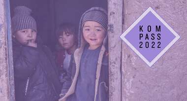 Drei Kinder in Afghanistan stehen in einer Tür und schauen Richtung Kamera. Rechts oben liegt eine Grafik über dem Foto mit dem violetten Logo des Kompass 2022.