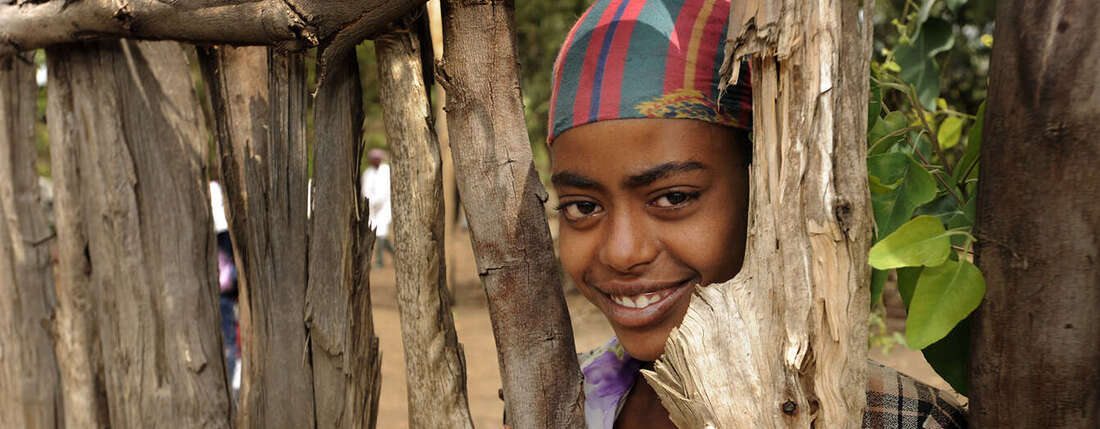 Kind schaut zwischen Baumstämmen hindurch und lacht in die Kamera.