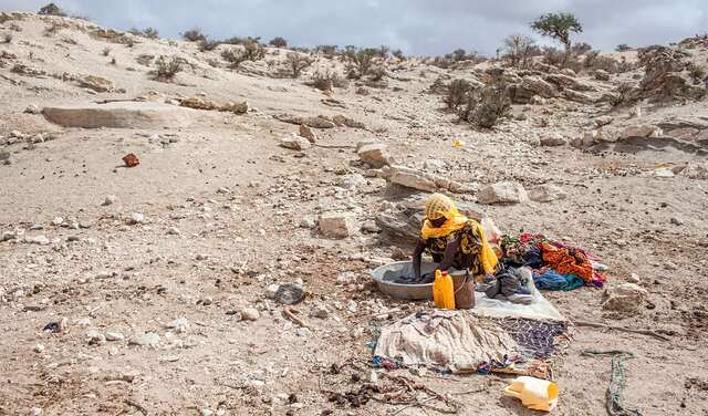 Eine Frau wäscht in einem Bottich Wäsche, im Hintergrund ist eine karge Landschaft zu sehen.