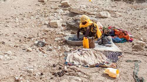 Eine Frau wäscht in einem Bottich Wäsche, im Hintergrund ist eine karge Landschaft zu sehen.