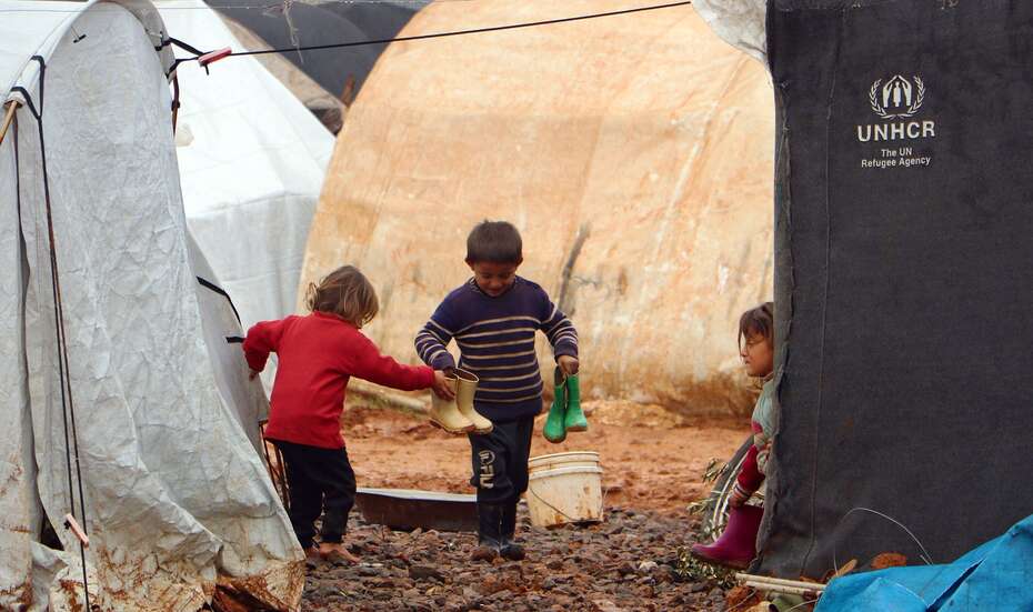 Drei Kinder stehen zwischen Zelten in einem Flüchtlingscamp. Sie halten Gummistiefel in ihren Händen.
