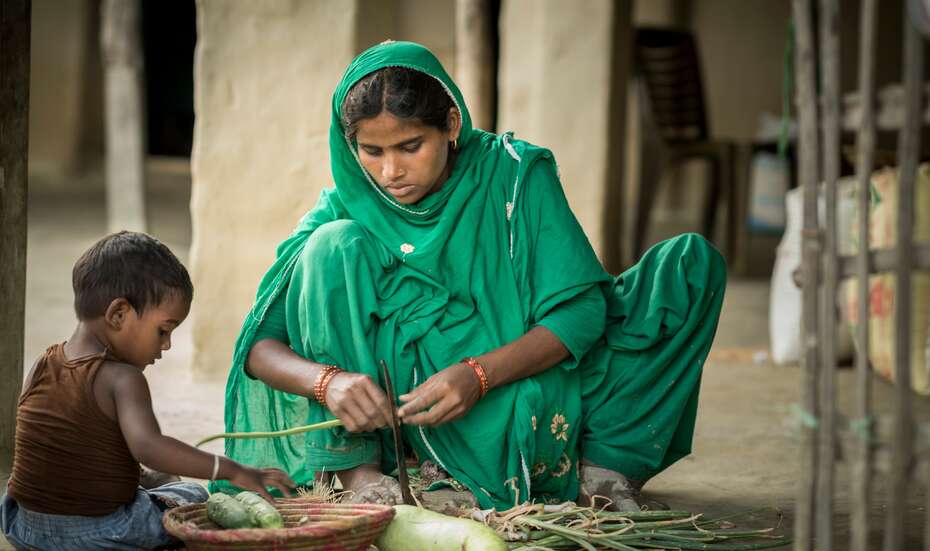 Eine Frau hockt auf dem Boden und bearbeitet Gemüse, neben ihr sitz ein Kind.