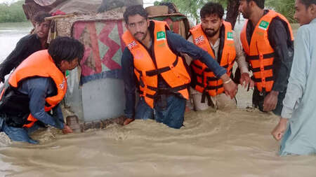 Überschwemmungen in Pakistan. Männer mit Schwimmwesten tragen einen großen Container durch kniehohes Wasser