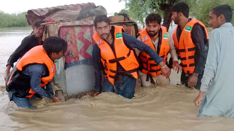 Überschwemmungen in Pakistan. Männer mit Schwimmwesten tragen einen großen Container durch kniehohes Wasser