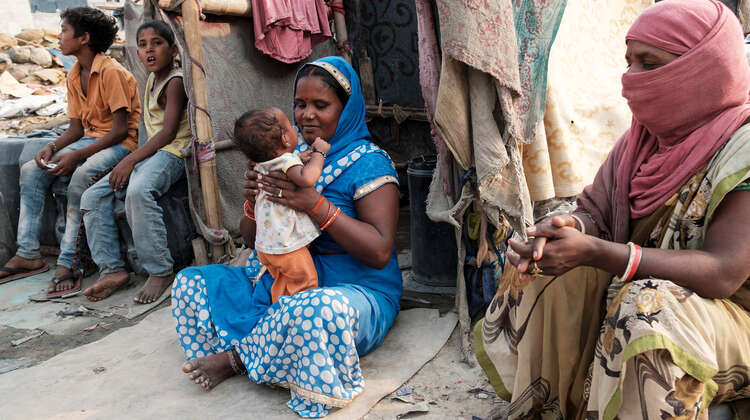 Frauen und Kinder sitzen auf dem Boden, im Hintergrund sieht man eine Notunterkunft.