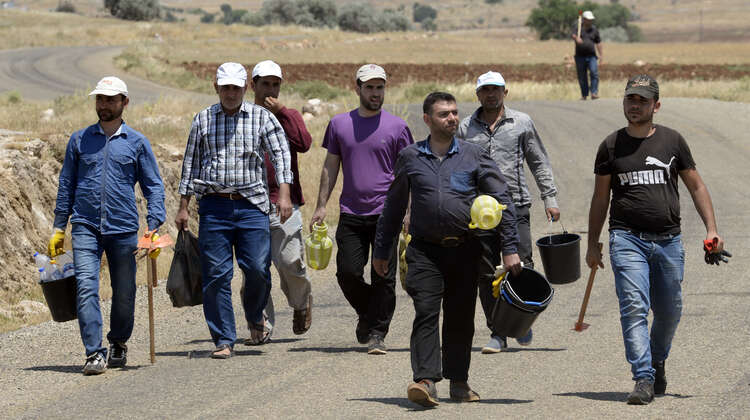 Acht Männer gehen eine Straße entlang. Sie tragen Eimer, Gießkannen und Werkzeuge. 
