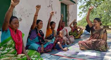 Frauen in Indien, draußen auf dem Boden sitzend bei einem Beratungsangebot. Sie strecken die Faust Richtung Himmel.