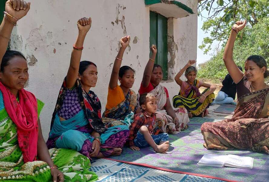 Frauen in Indien, draußen auf dem Boden sitzend bei einem Beratungsangebot. Sie strecken die Faust Richtung Himmel.