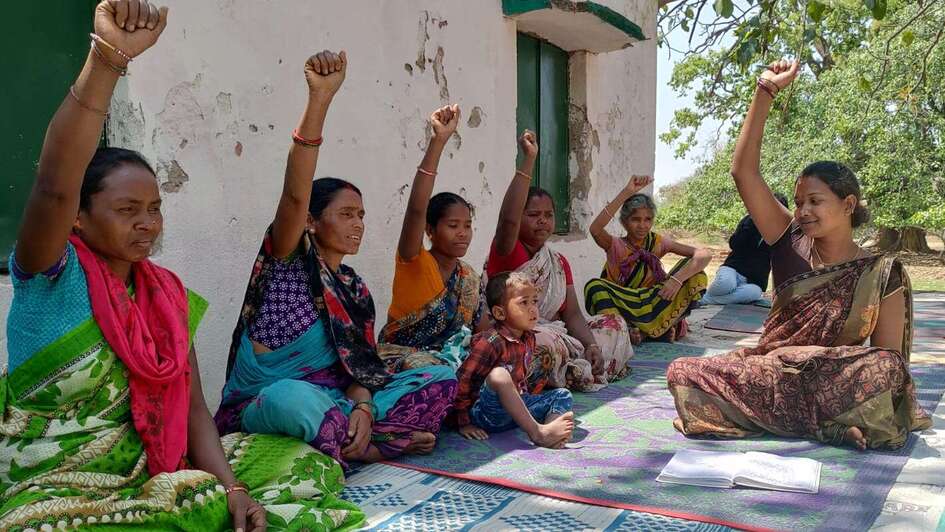 Frauen in Indien, draußen auf dem Boden sitzend bei einem Beratungsangebot. Sie strecken die Faust Richtung Himmel.