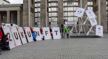 Aktionsgruppe vor dem Brandenburger Tor vor einem symbolisch einstürzenden Kartenhaus. Sie halten große Karten mit der Aufschrift "Zukunft".