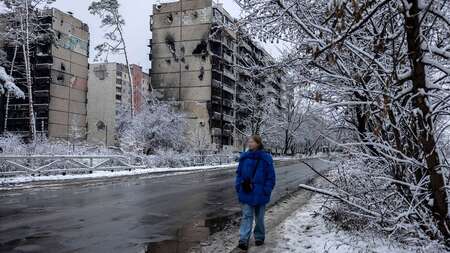 Eine Frau läuft eine Straße entlang, im Hintergrund zerstörte Wohnhäuser, es ist kalt und verschneit