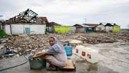 Eine Frau sitht auf einem Stück Holz, vor ihr ein Wäschekübel. Um sie herum sieht man die Zerstörung, die der Tsunami angerichtet hat.