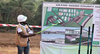 Ein Mann weist mit einem Zeigestock auf eine Tafel mit einem Bauplan.