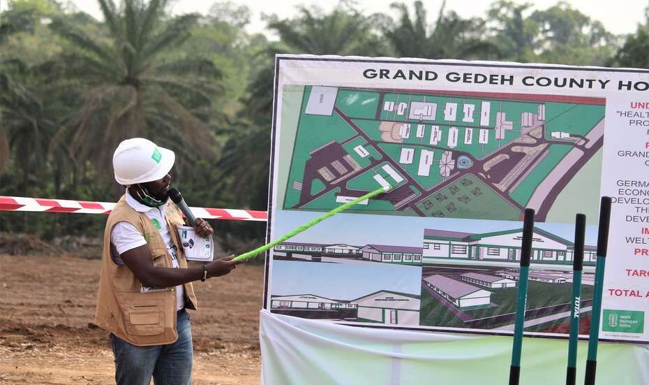 Ein Mann weist mit einem Zeigestock auf eine Tafel mit einem Bauplan.