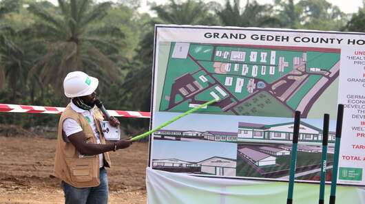 Ein Mann weist mit einem Zeigestock auf eine Tafel mit einem Bauplan.