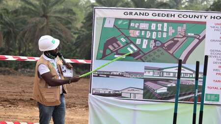 Ein Mann weist mit einem Zeigestock auf eine Tafel mit einem Bauplan.