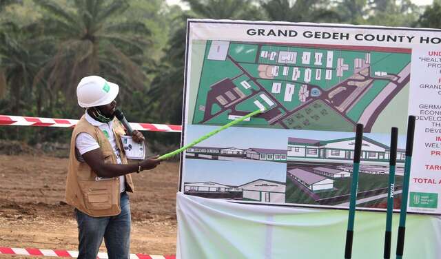 Ein Mann weist mit einem Zeigestock auf eine Tafel mit einem Bauplan.