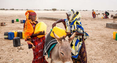 Zwei junge Frauen laden mit Wasser gefüllte Kanister auf einen Esel, im Hintergrund ist eine trockene Landschaft zu sehen.