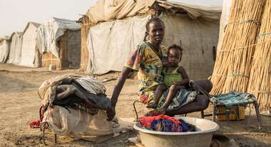 Eine Frau sitzt mit ihrem Baby auf dem Schoß vor einem Waschzuber voller Wäsche.