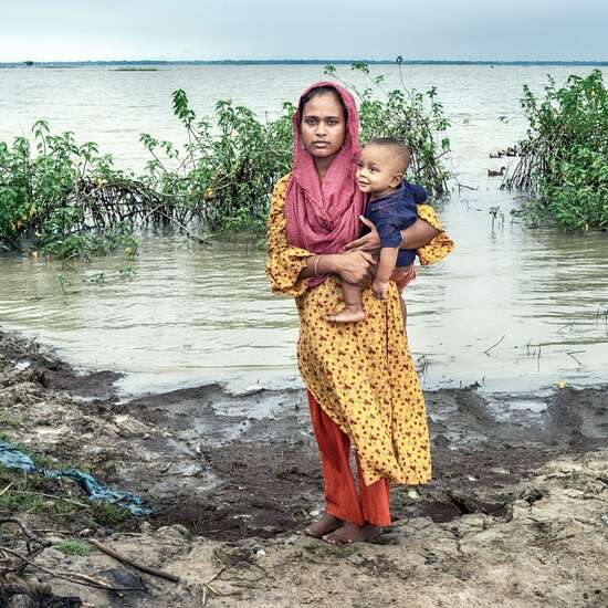 Eine Mutter mit ihrem Kind auf dem Arm steht vor überschwemmtem Land in Bangladesch.