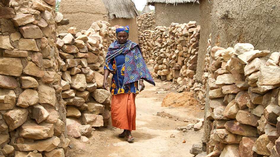 Eine Frau in Mali läuft zwischen Lehmhütten entlang.