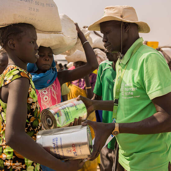 Im Vordergrund: Eine Frau erhält von einem Mann in grünem T-shirt Nahrungsmittel. Im Hintergrund sieht man Menschen, die in einer Schlange anstehen.