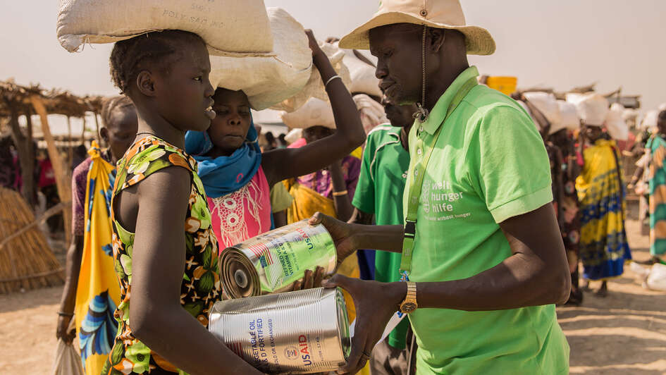 Im Vordergrund: Eine Frau erhält von einem Mann in grünem T-shirt Nahrungsmittel. Im Hintergrund sieht man Menschen, die in einer Schlange anstehen.