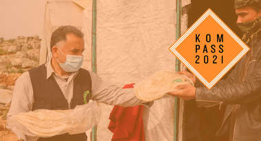 Ein Mann händigt einem anderen Mann eine Tüte mit Fladenbrot. Oben rechts ist das Kompass 2021 Logo.